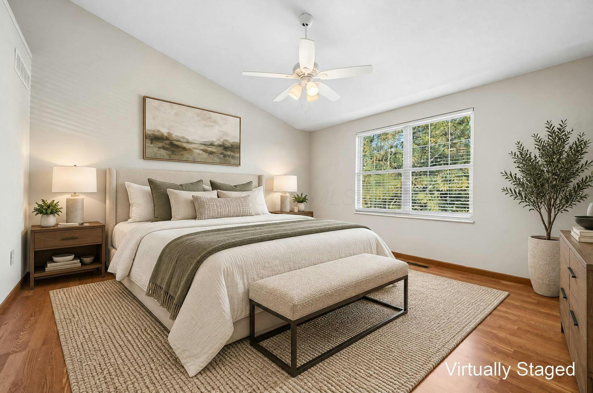Virtually staged bedroom with a large neutral bed, hardwood floors, and a wide window overlooking trees.