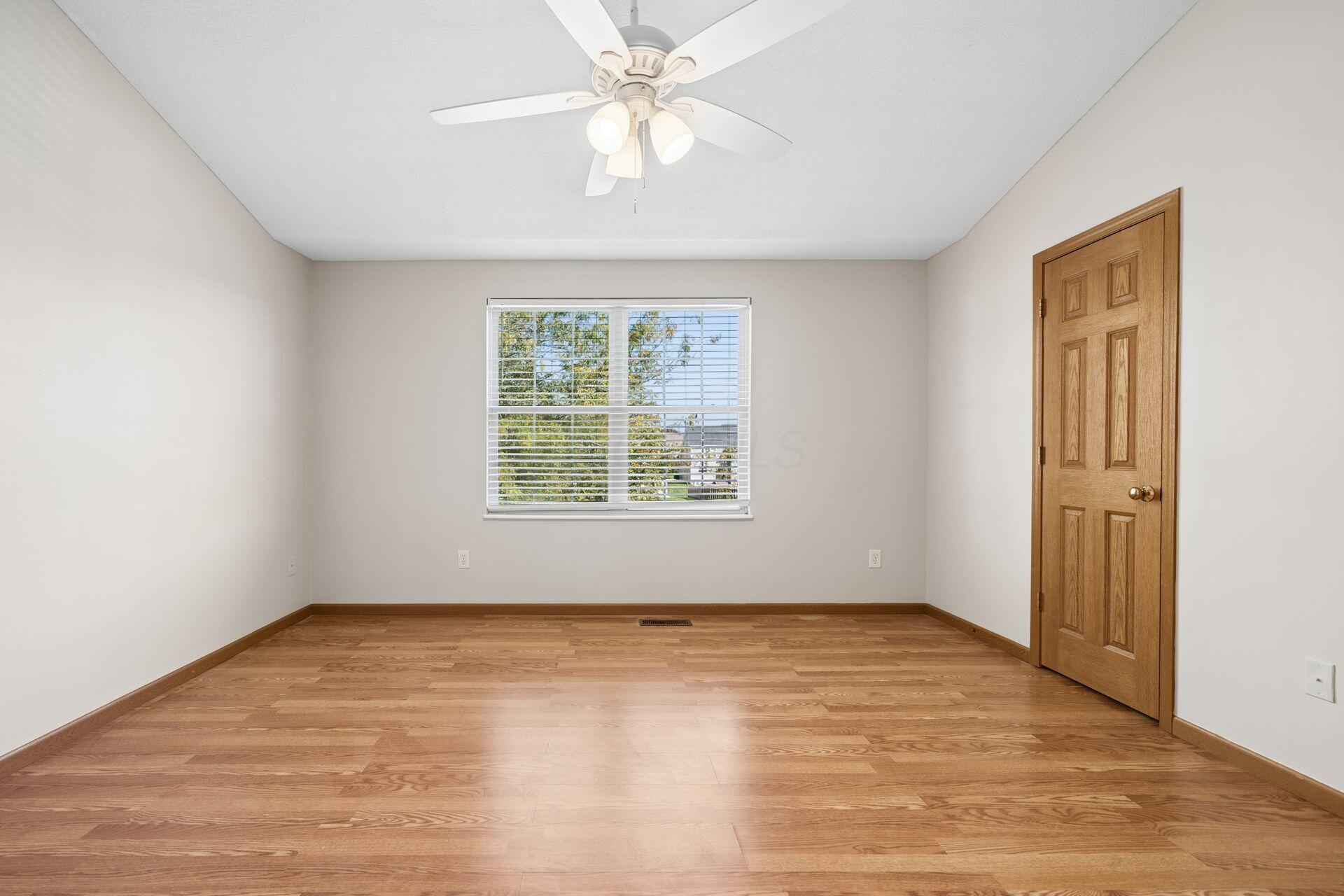 Empty bedroom with hardwood floors, a ceiling fan, and a window with white blinds.