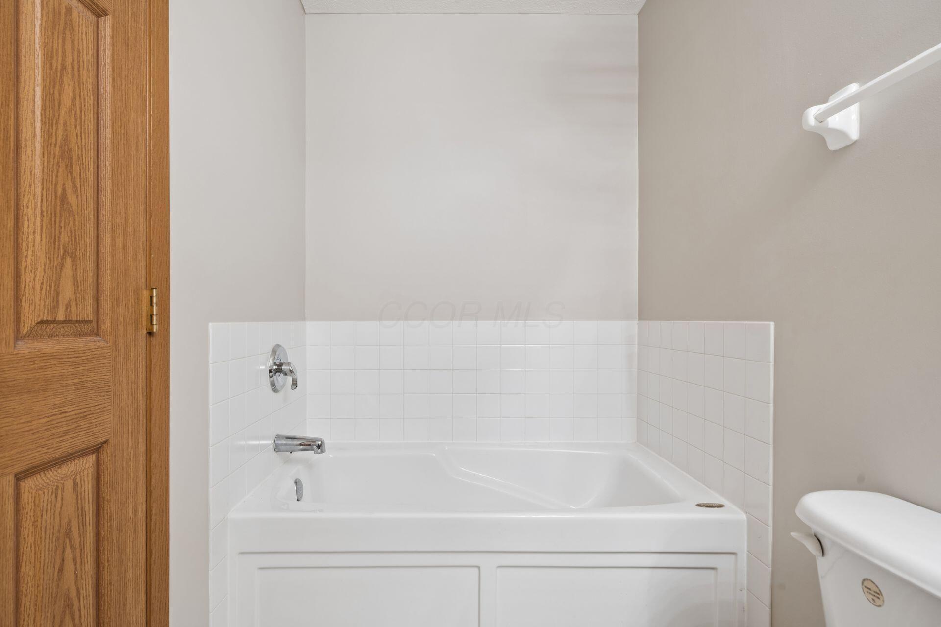 Bathroom bathtub with white tiled surround and chrome fixtures beside a wooden door.