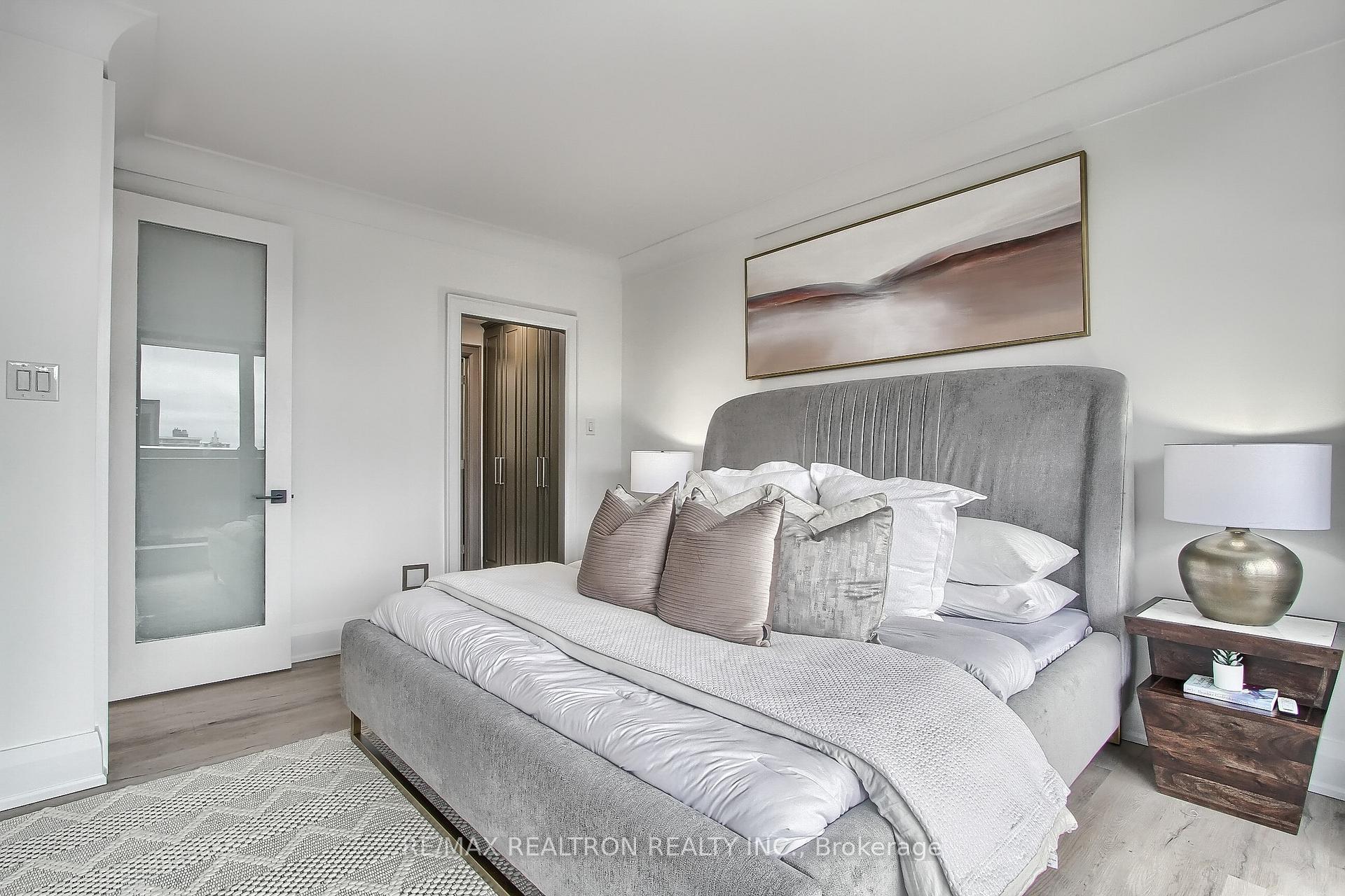Staged bedroom with a gray upholstered bed, layered white bedding, abstract art, and a geometric rug.