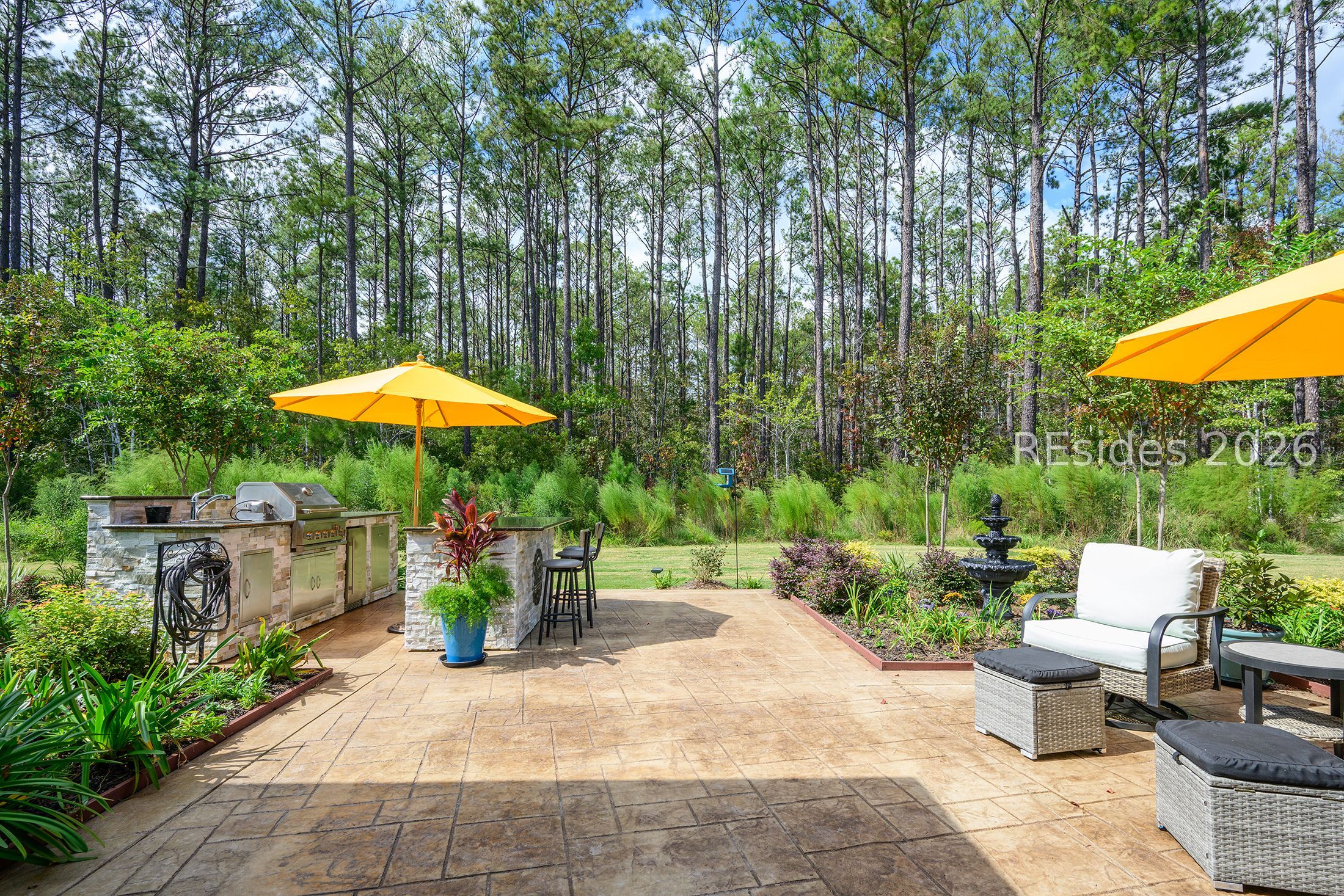 Outdoor patio with a stone kitchen, yellow umbrellas, lounge seating, and a wooded backdrop.