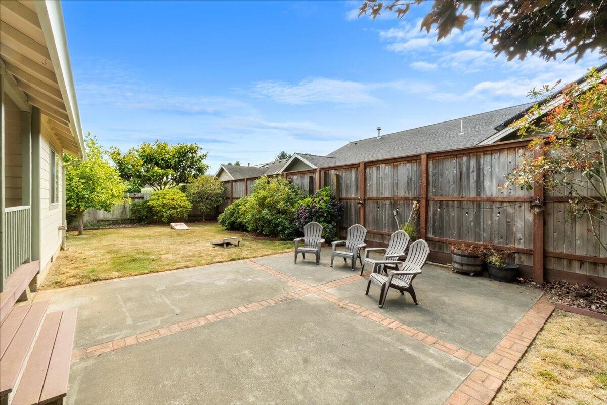 Fenced backyard patio with Adirondack chairs, brick edging, a small lawn, and shrubs.