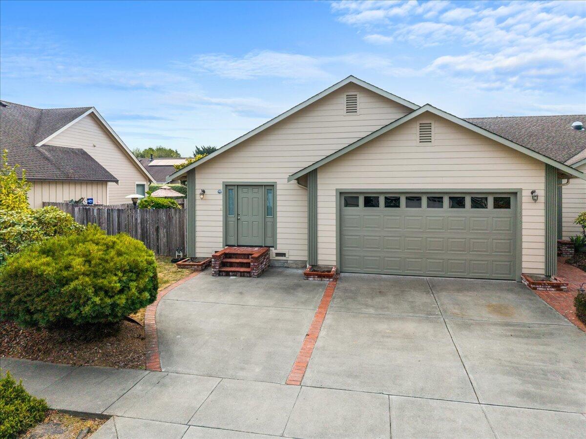 Single-story beige home with sage-green trim, a two-car garage, and a brick-edged driveway.