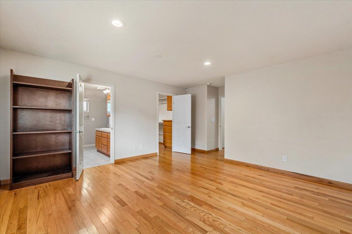 Empty bedroom with hardwood floors and a tall bookshelf, with the bathroom visible through an open door.