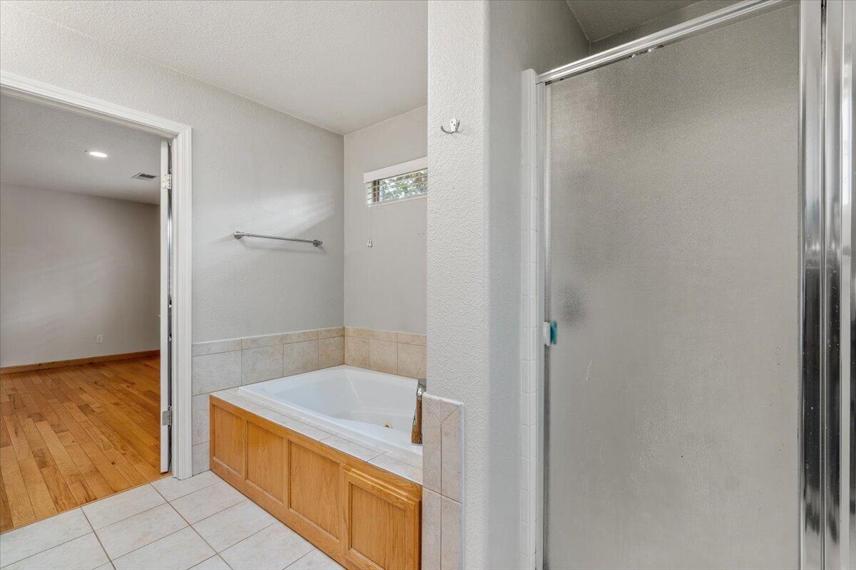 Bathroom with a tiled soaking tub and frosted-glass shower beside a doorway to a hardwood-floored room.
