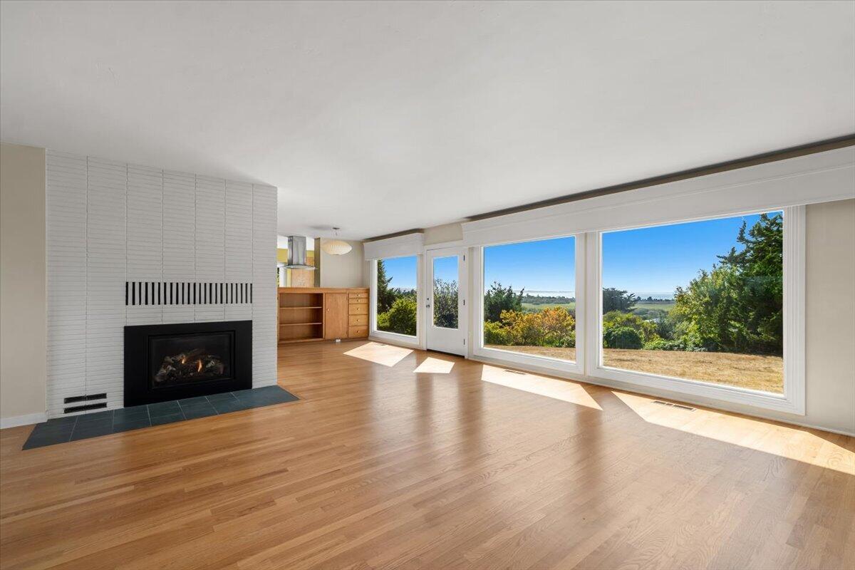 Vacant living room with a tiled fireplace, hardwood floors, and large windows overlooking greenery.