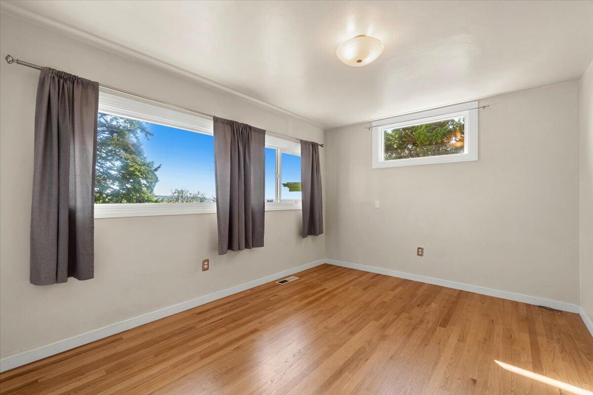 Empty room with hardwood floors and curtained windows overlooking trees.