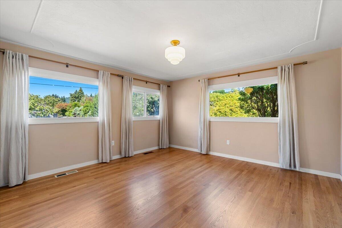 Empty room with wood floors and three curtained windows overlooking trees.