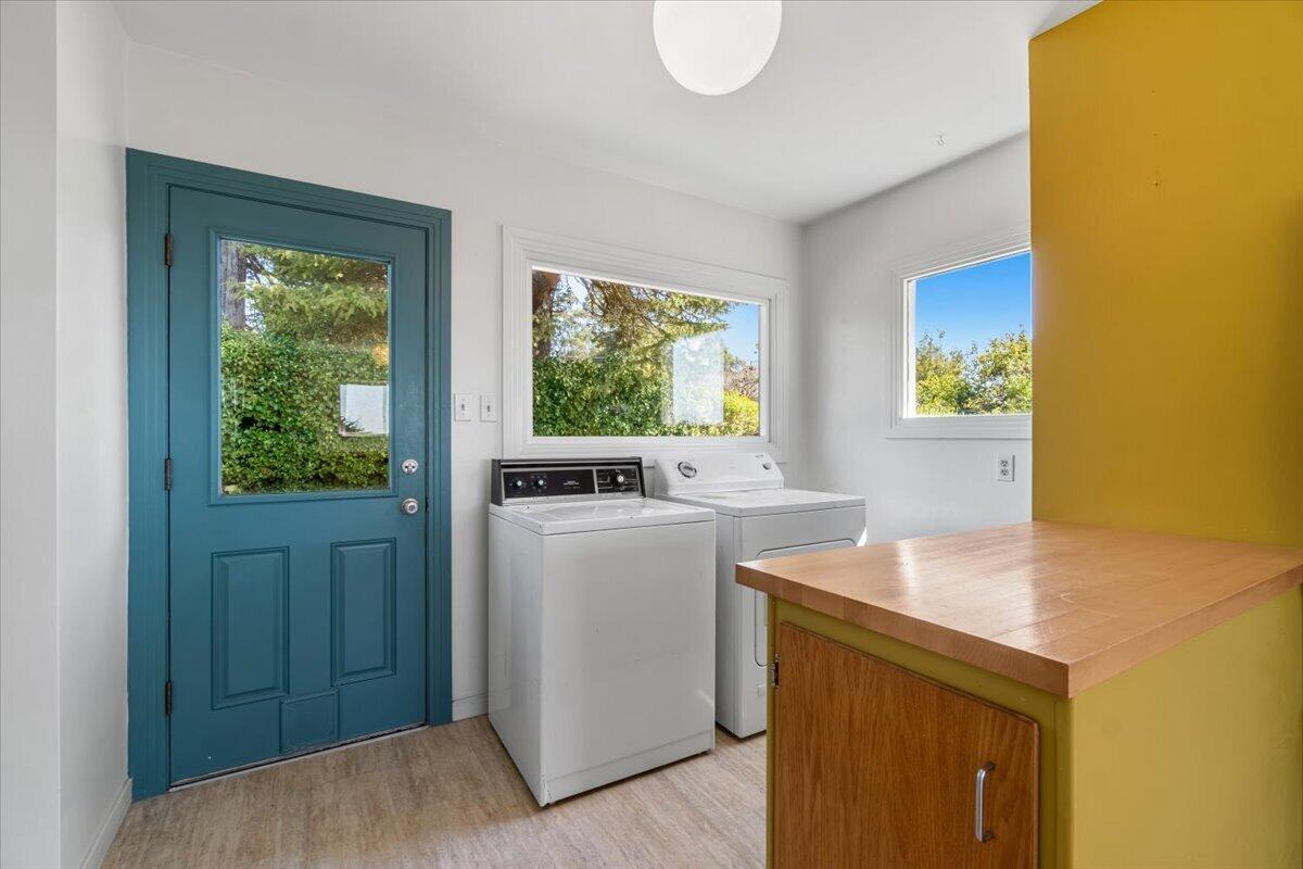Laundry room with a teal door, two white machines, garden-facing windows, and a wood-topped yellow cabinet.