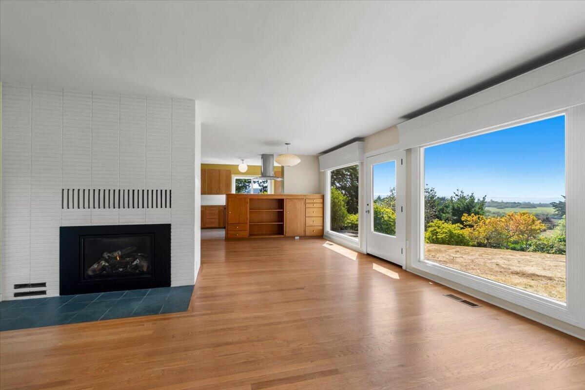 Vacant living room with a white tile fireplace, built-in wooden cabinets, and large windows overlooking trees.