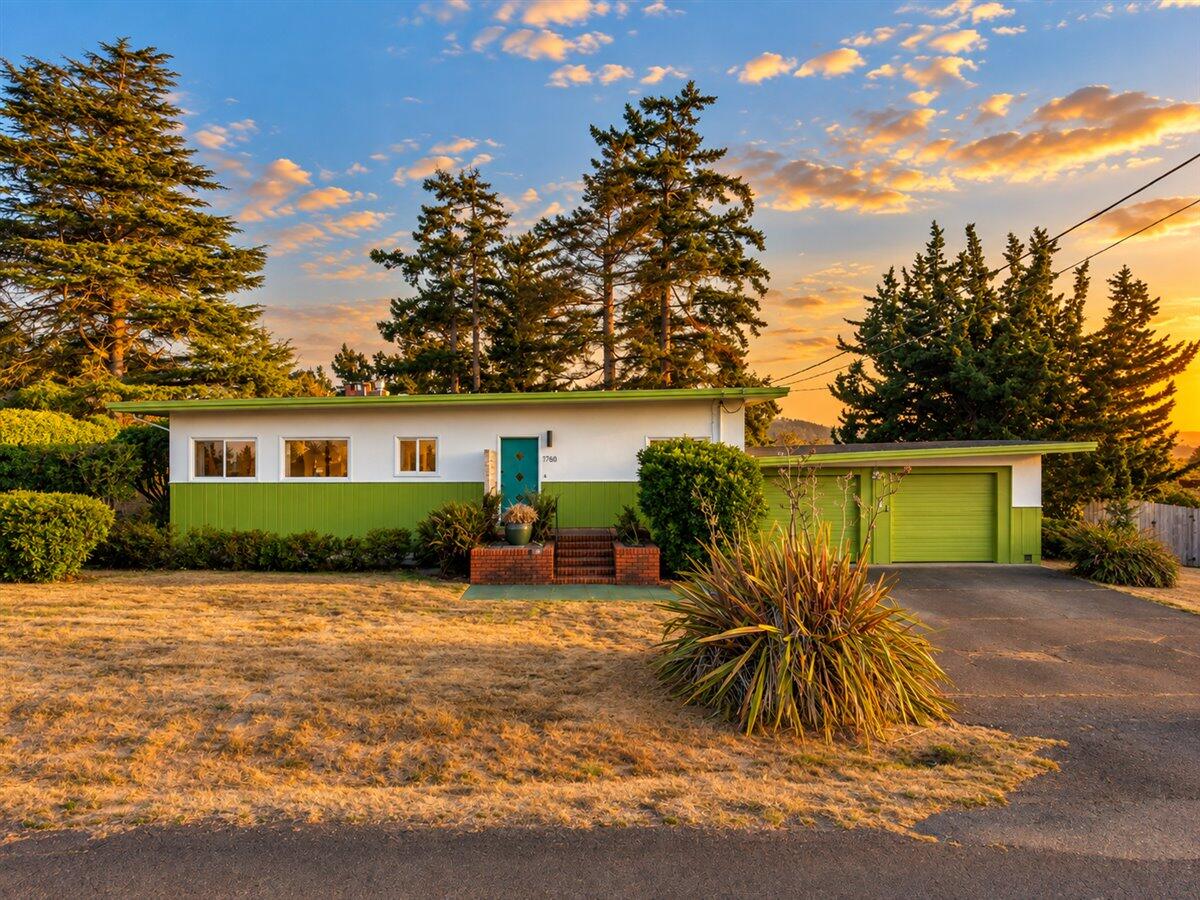 Single-story white-and-green home with a matching garage, framed by tall evergreens at sunset.