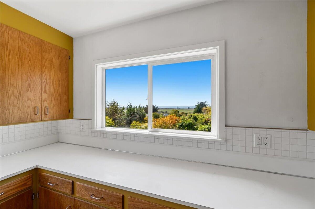 Kitchen with wood cabinets and white counters, looking through a large window toward the ocean.