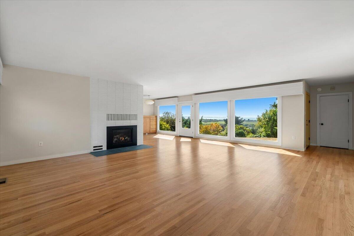 Vacant living room with hardwood floors, a tiled fireplace, and broad windows overlooking greenery.
