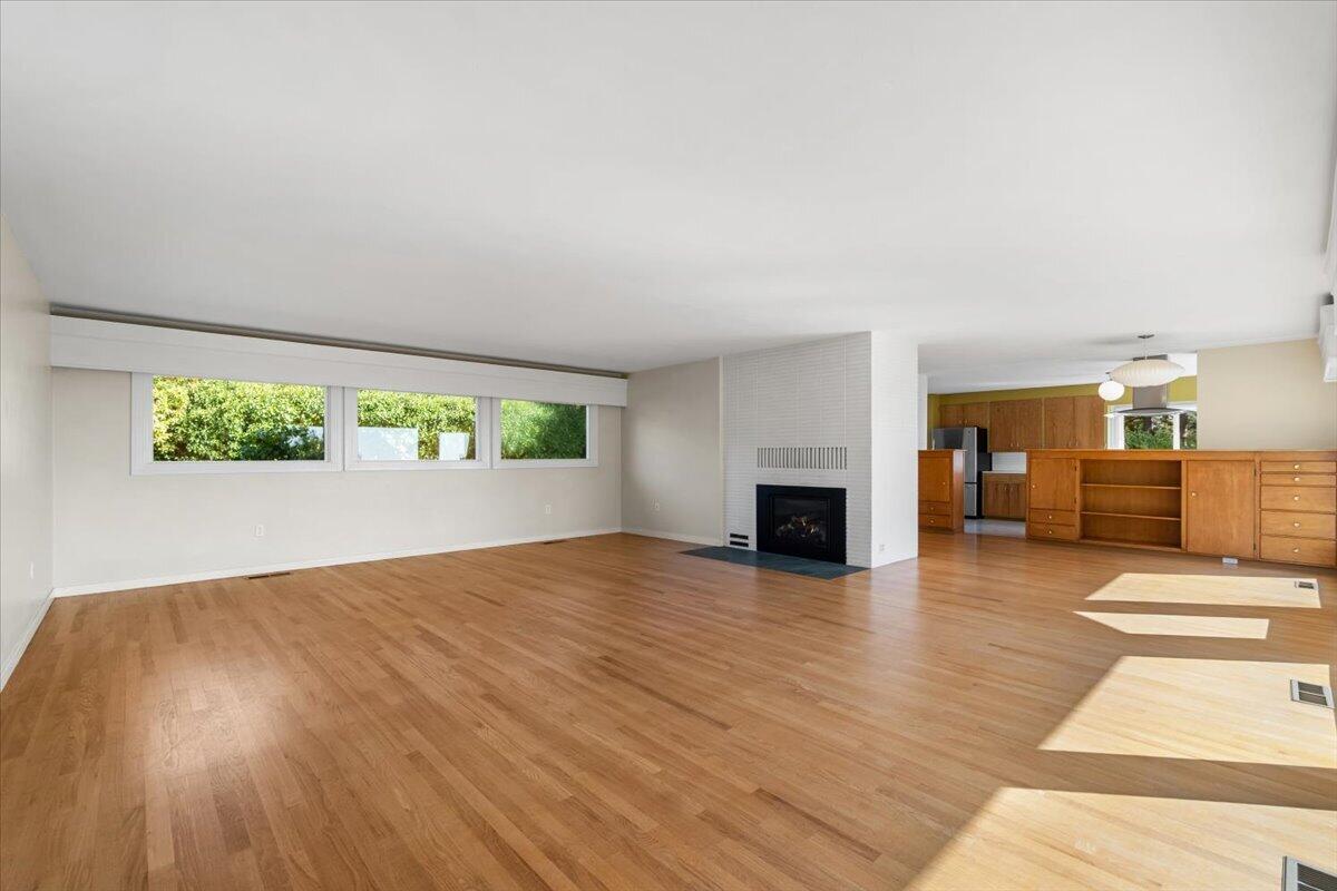 Vacant living room with hardwood floors, a tiled fireplace, and windows opening to the kitchen.