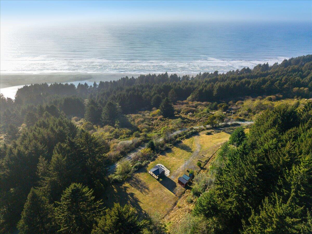 Aerial coastal property with a forested clearing, small outbuildings, and oceanfront beach.