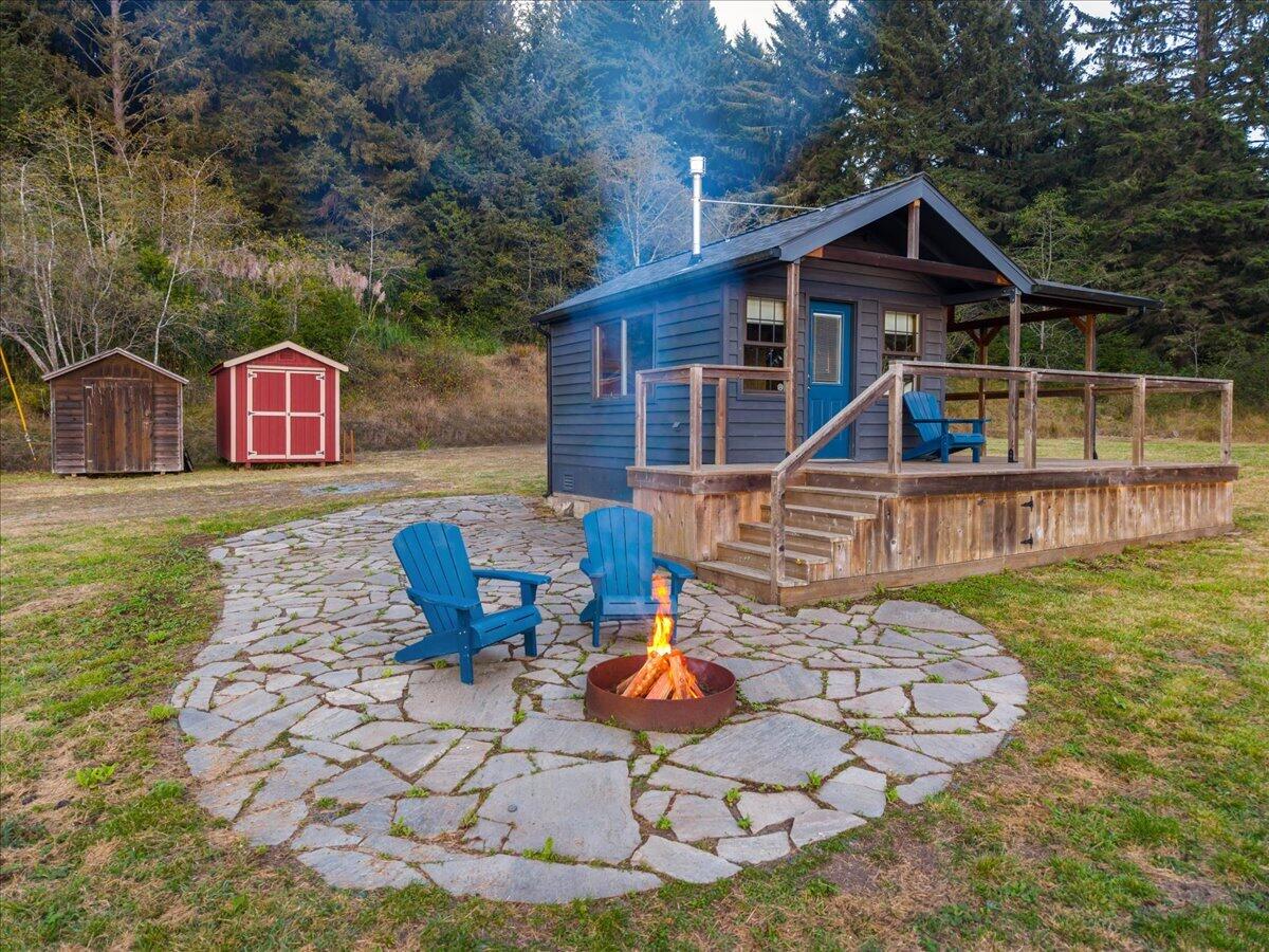 Blue cabin with a raised deck, stone patio, fire pit, Adirondack chairs, and two sheds.