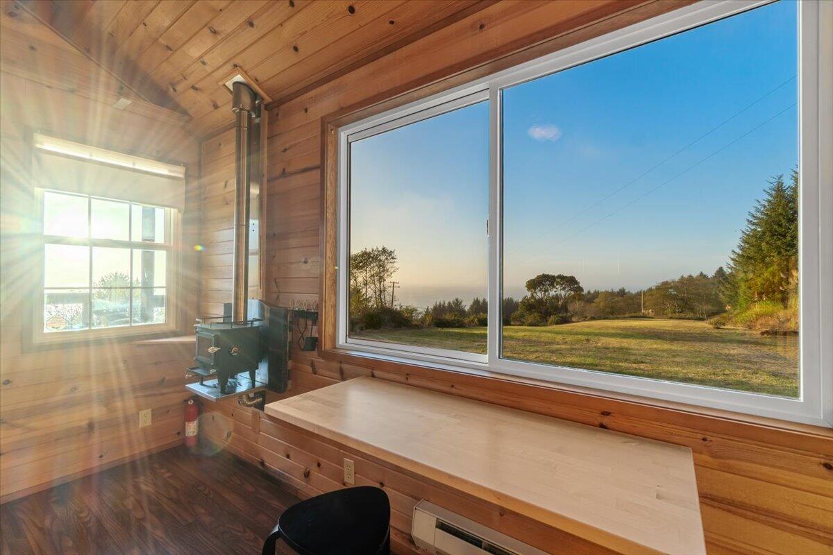 Wood-paneled room with a wood stove and broad window overlooking a grassy clearing and distant coast.