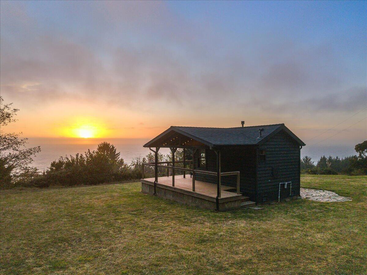 Dark coastal cabin with a covered deck overlooking the ocean at sunset.