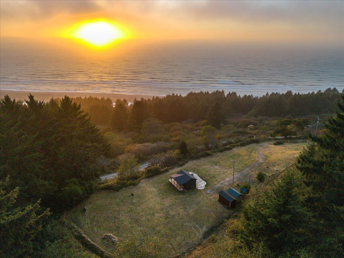 Aerial view of a coastal clearing with two sheds and the Pacific sunset beyond the trees.