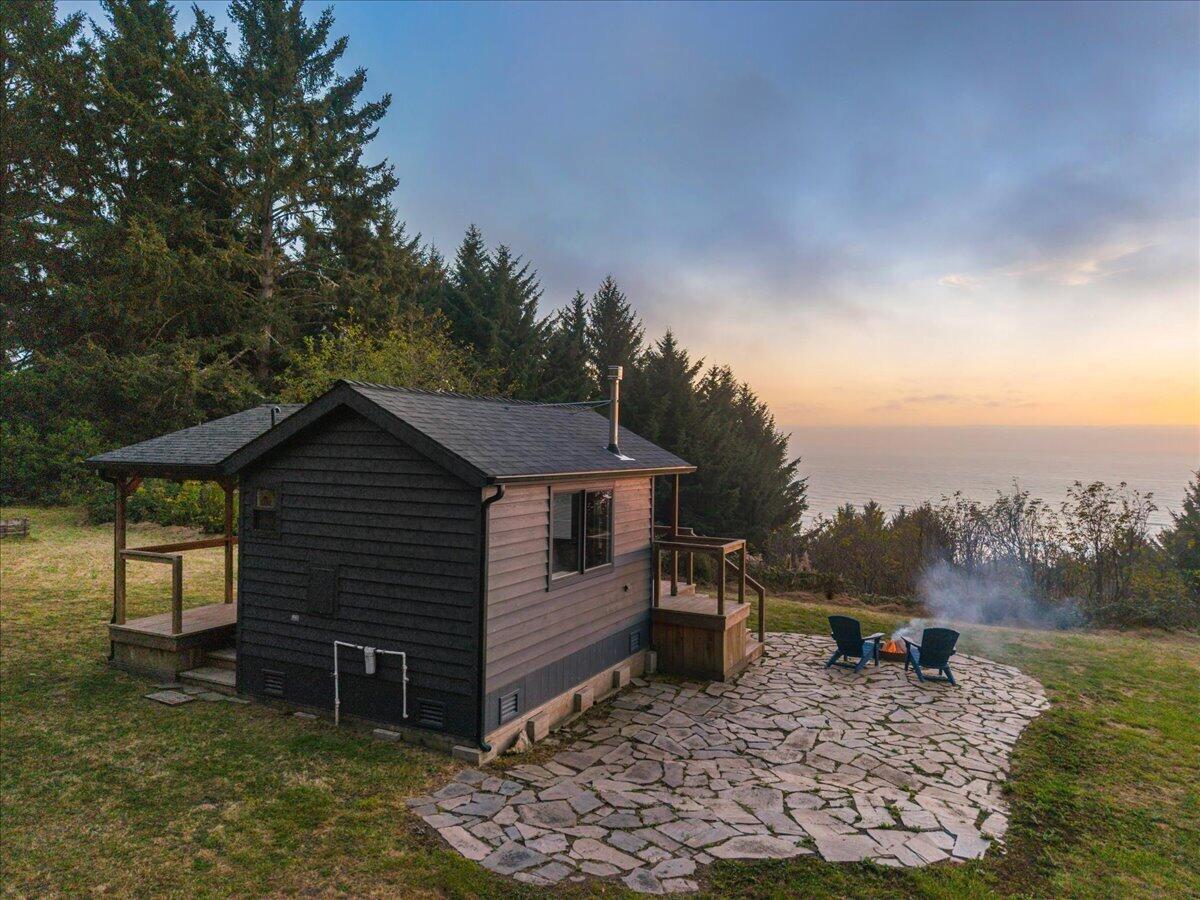 Small coastal cabin beside a stone patio, with trees and ocean views at sunset.