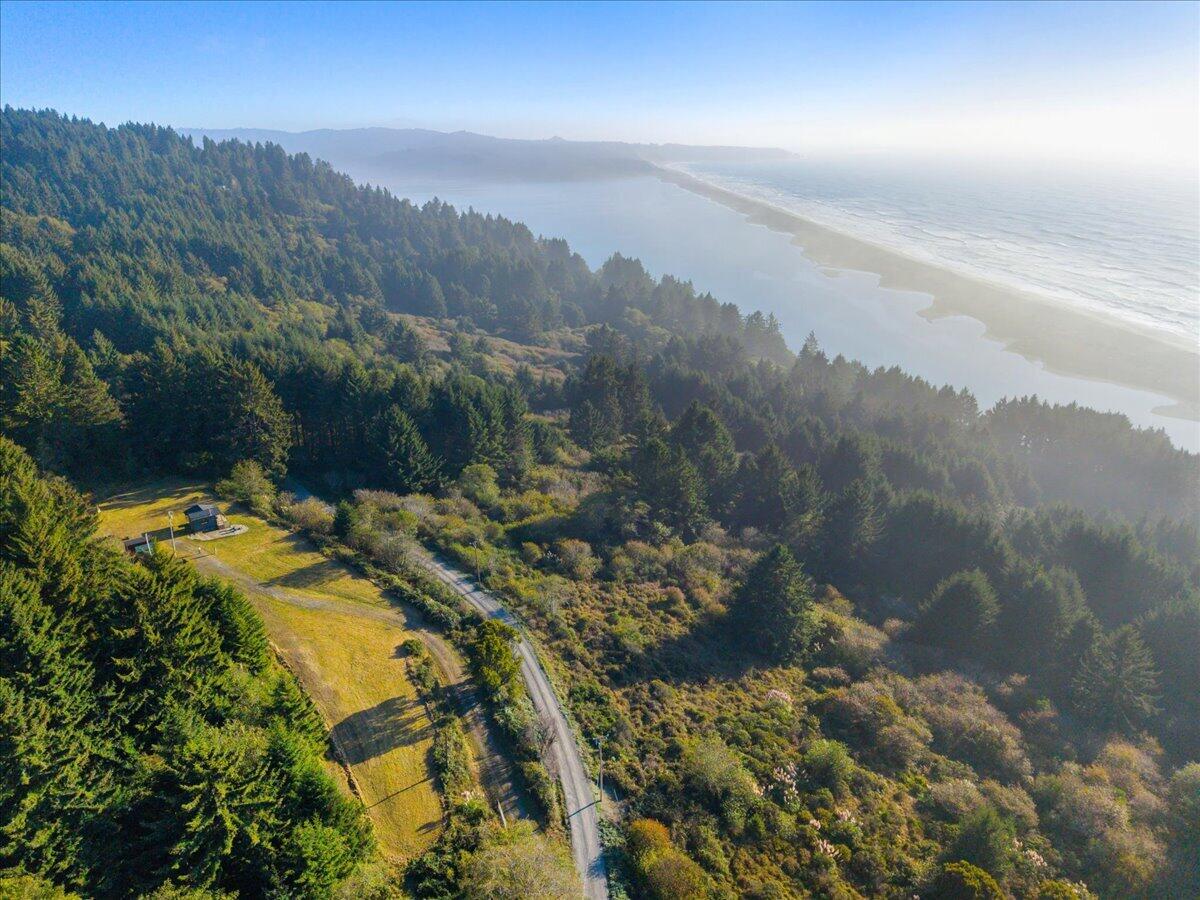 Aerial coastal property with a small home in a forest clearing beside a winding road, ocean, and beach.