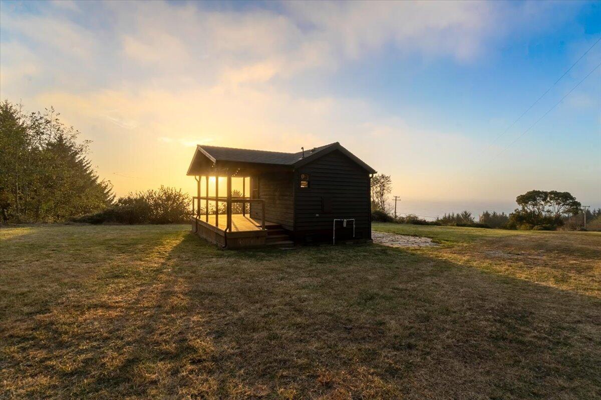 Dark wood cabin with a covered porch and deck in a grassy clearing, ocean visible at sunset.