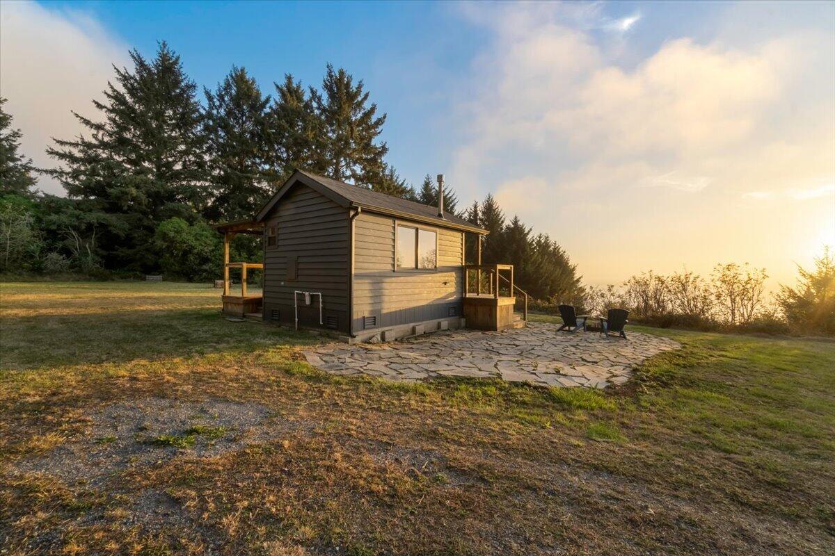 Small cabin with a stone patio and outdoor chairs on a grassy, tree-lined lot at golden hour.