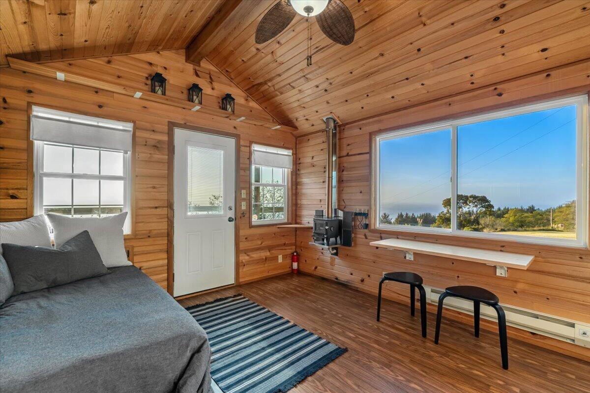 Wood-paneled cabin bedroom with a gray bed, black wood stove, and ocean view through a large window.