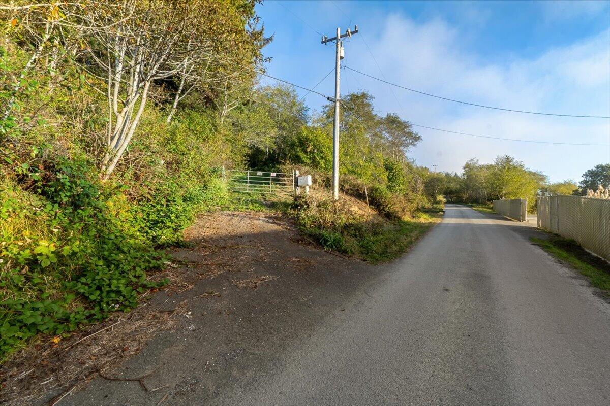 Gated gravel driveway entrance beside a paved road, bordered by dense trees and brush.