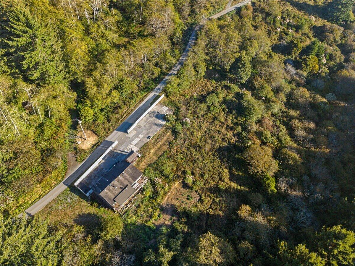Aerial view of a home and outbuildings amid wooded land beside a narrow road.