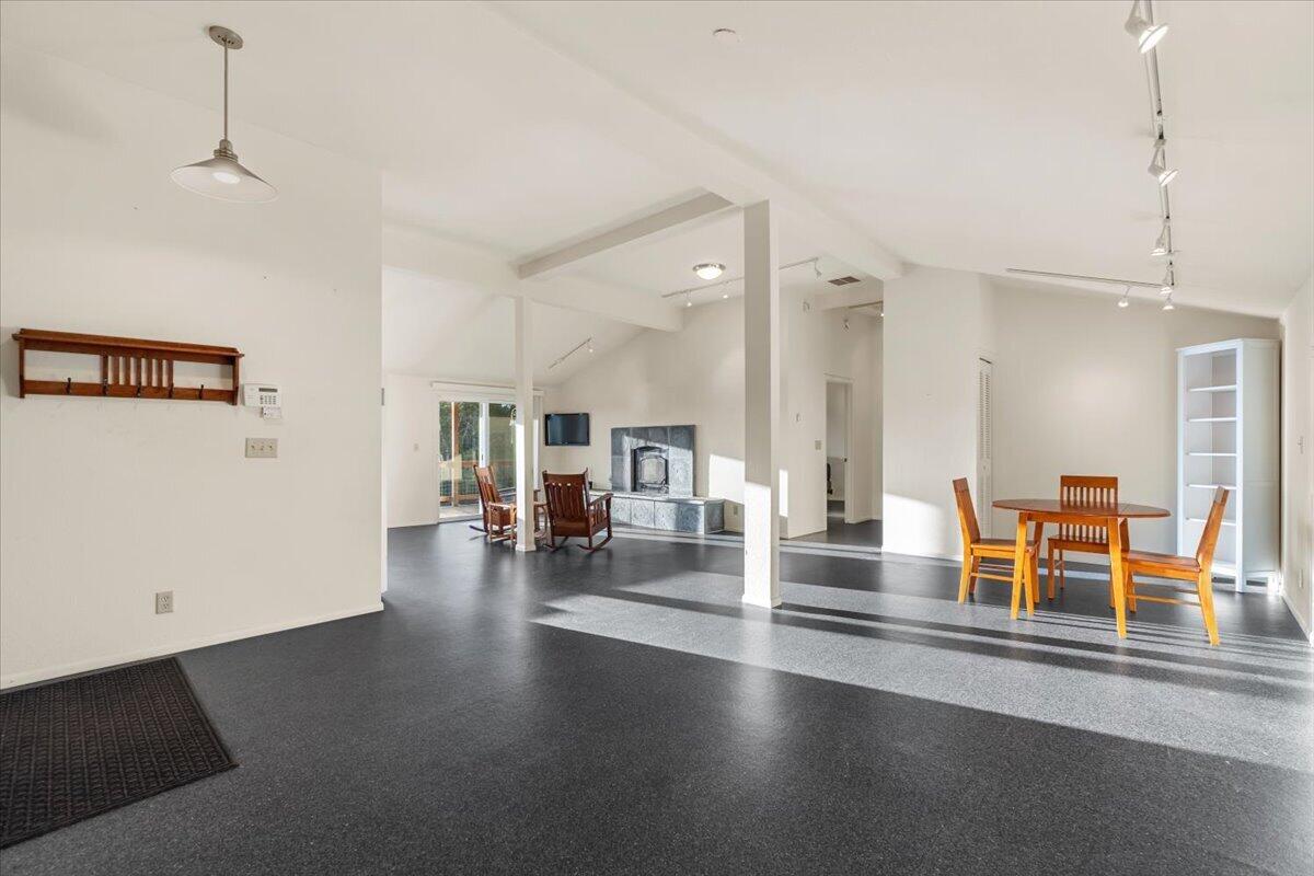Open-plan living and dining area with a stone fireplace and sunlight across dark floors.