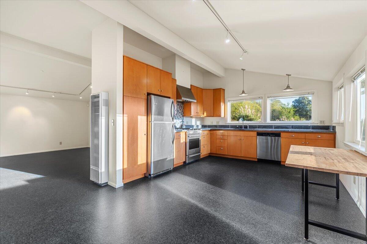 Open kitchen with warm wood cabinets, stainless steel appliances, and a wooden table by large windows.