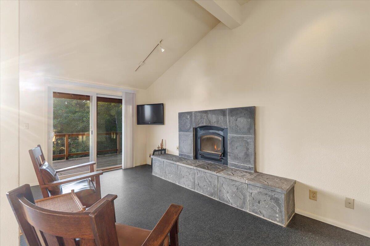 Living room with a stone fireplace, wooden chairs, and sliding doors to a tree-lined deck.