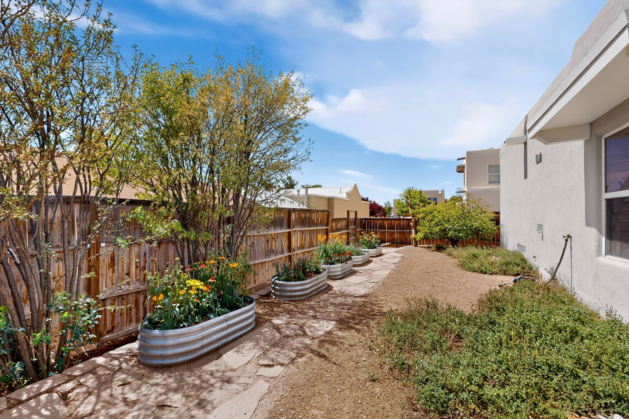 Side yard with a stone path and flower-filled raised garden beds along a wooden fence.
