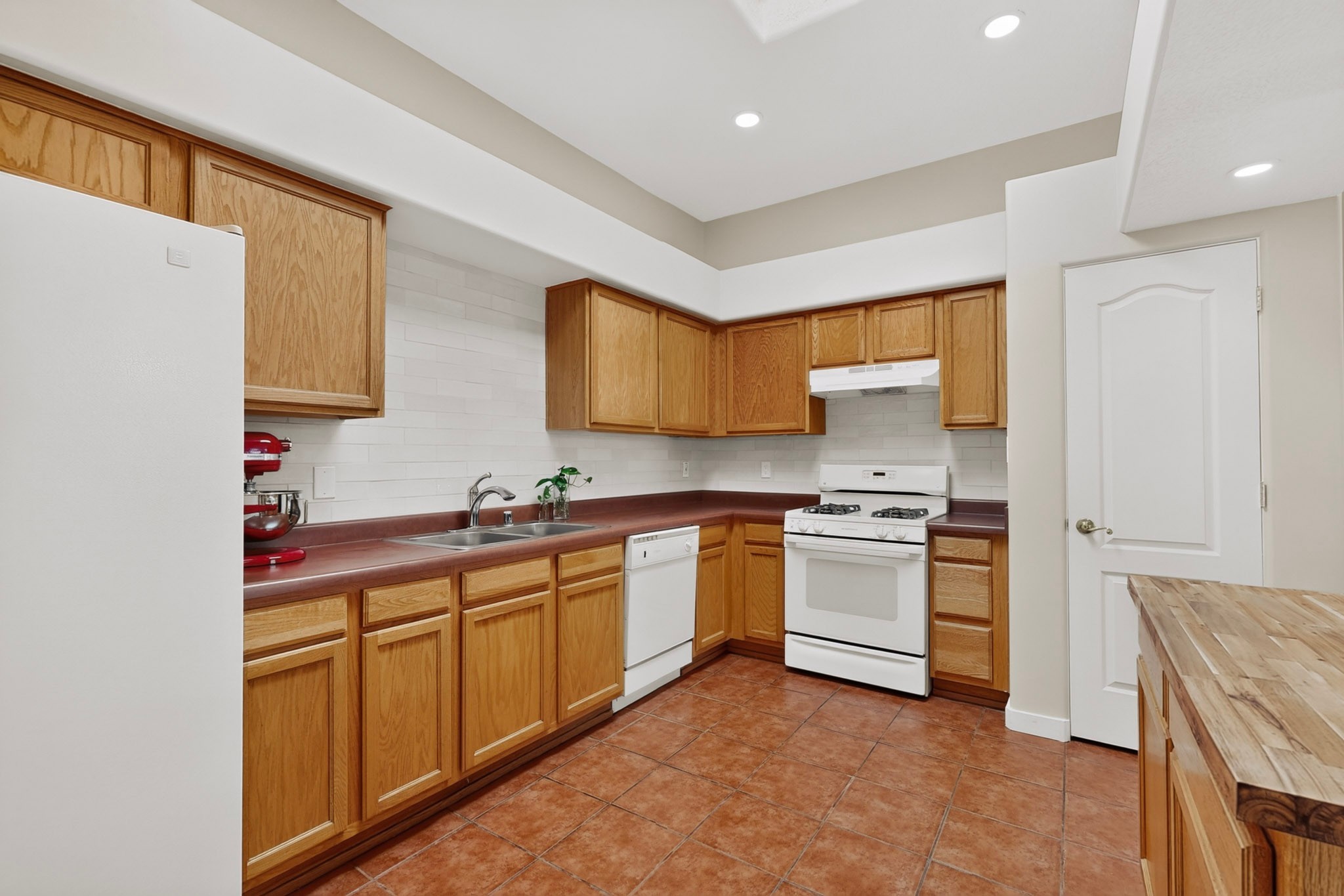 Kitchen with honey-colored wood cabinets, reddish-brown countertops, white appliances, and terracotta floor tiles.