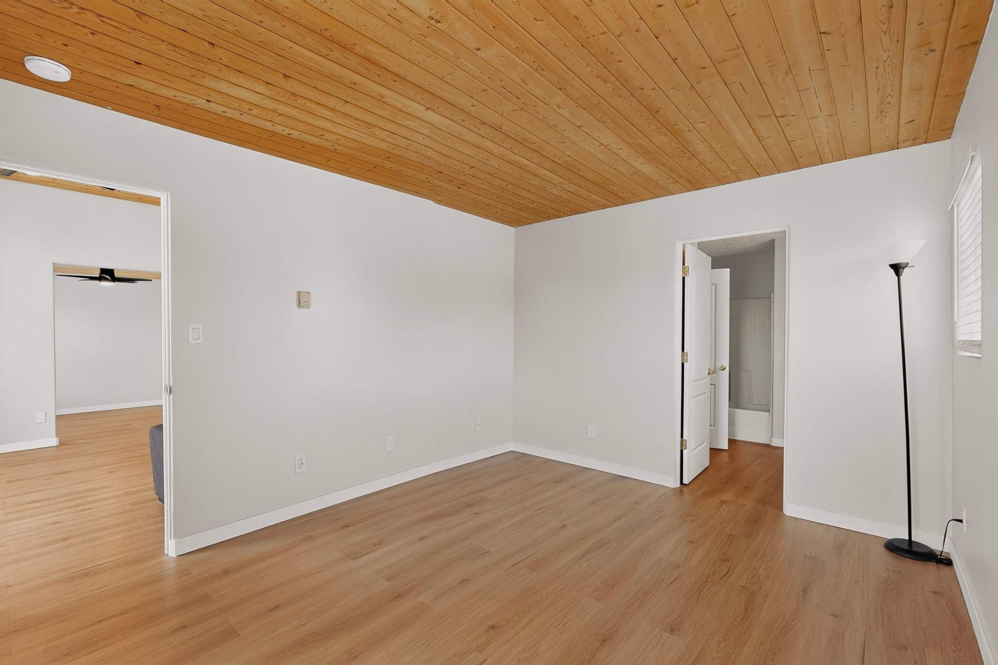 Empty room with a wood plank ceiling and floors, a floor lamp, and an open bathroom doorway.