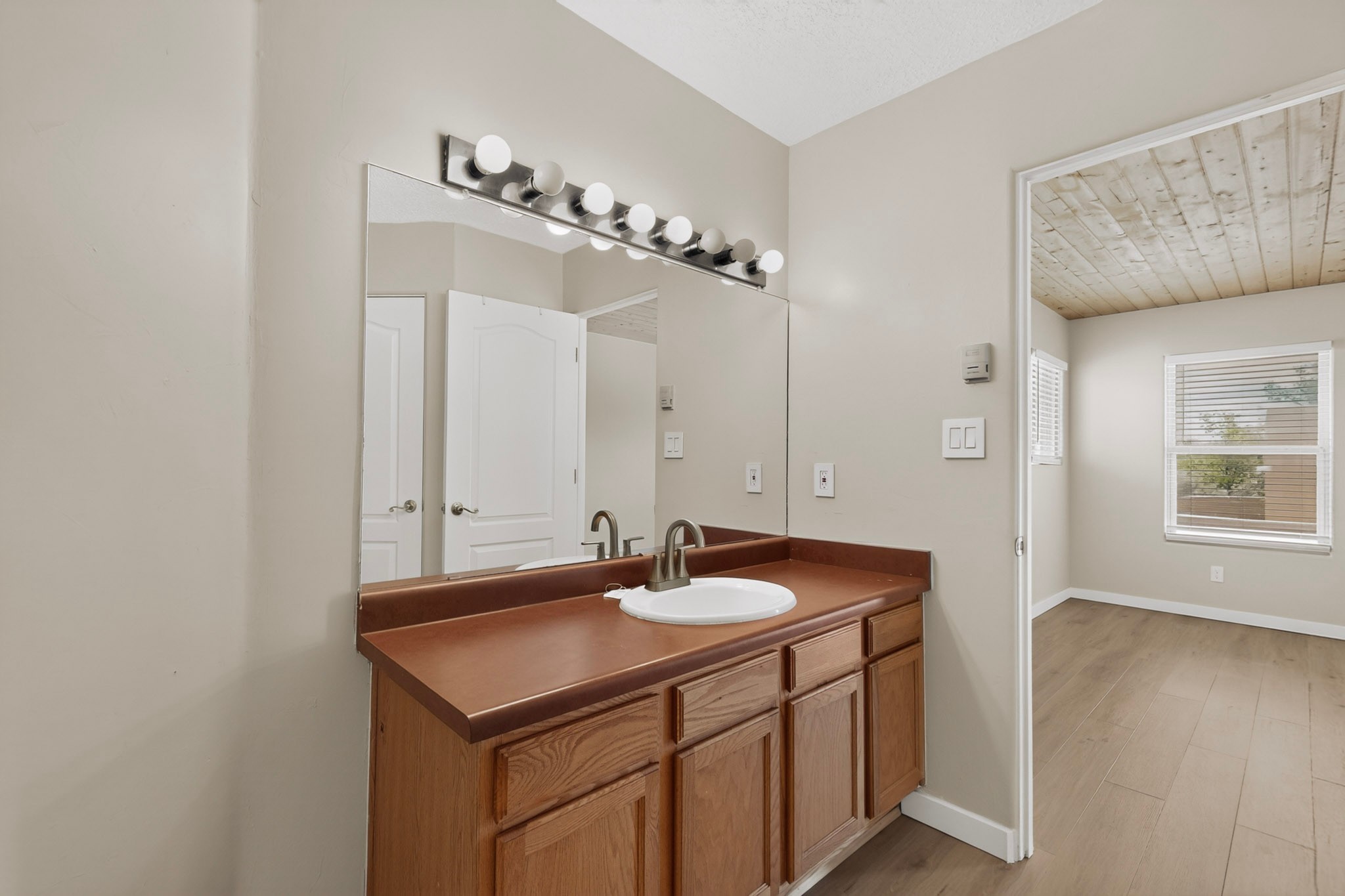 Bathroom vanity with a large mirror and globe lights, opening to a bright room with a wood-plank ceiling.