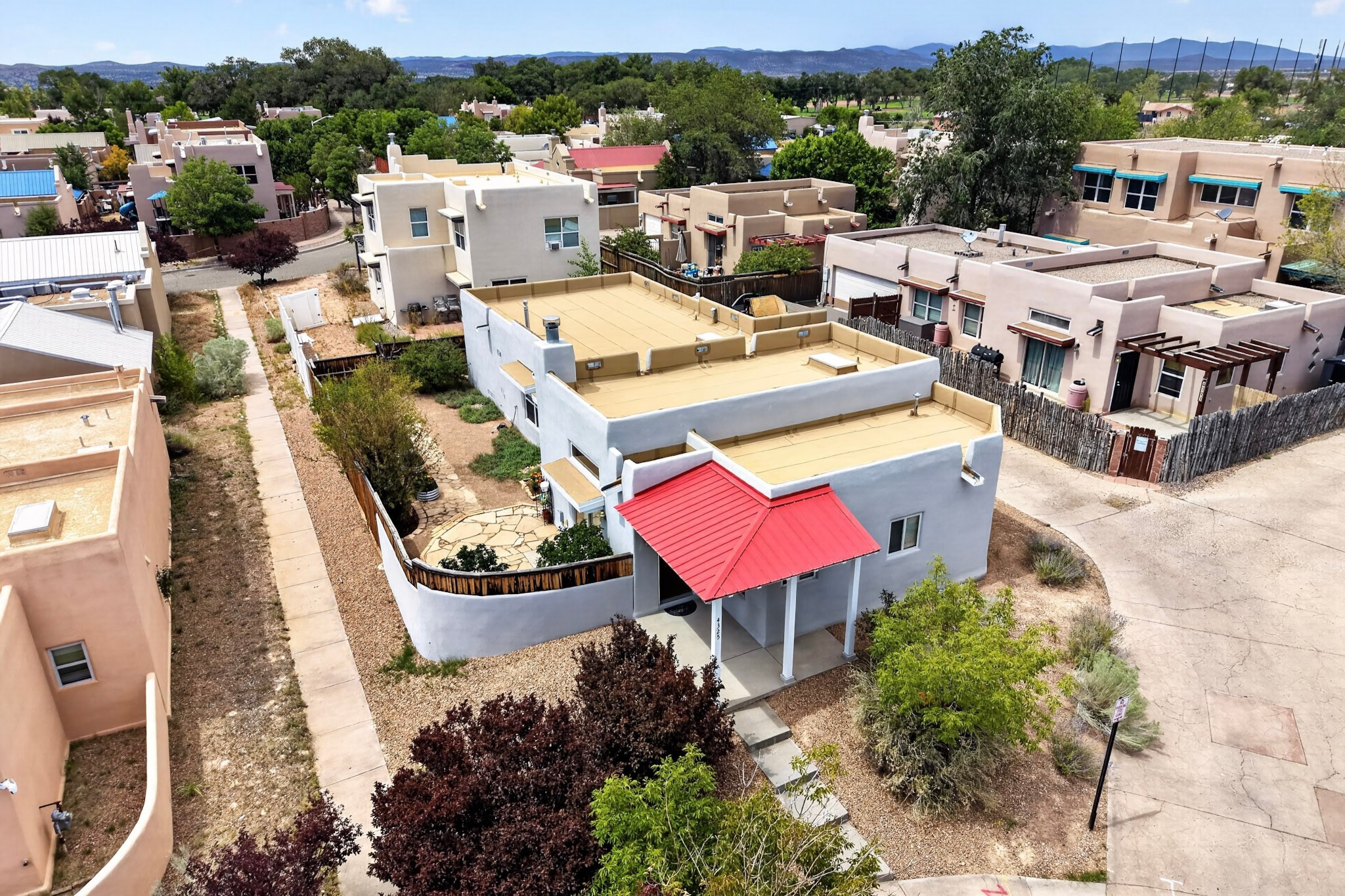 Aerial view of a gray stucco home with a red entry awning and flat rooftop, surrounded by neighborhood homes and distant mountains.