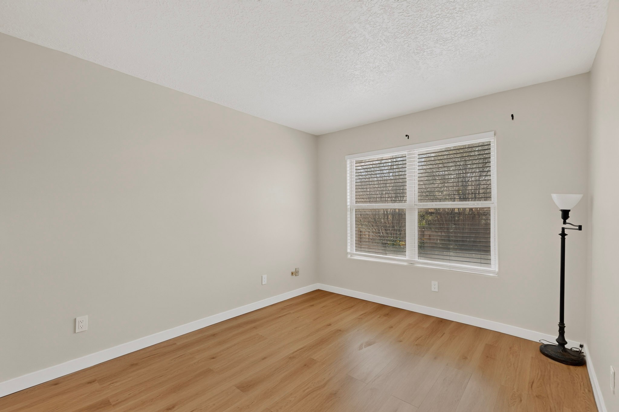 Empty room with light wood flooring, a large window with blinds, and a floor lamp.