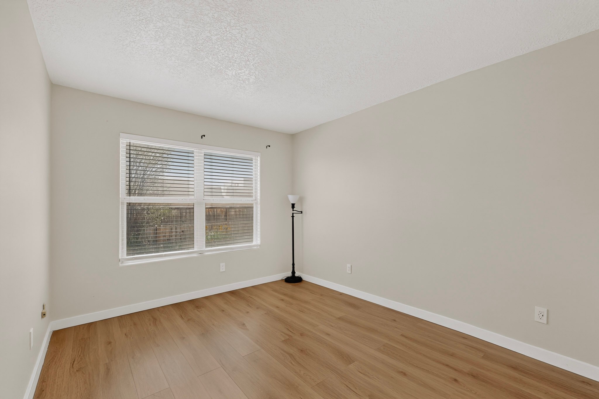 Empty room with wood flooring, a wide window with blinds, and a black floor lamp.
