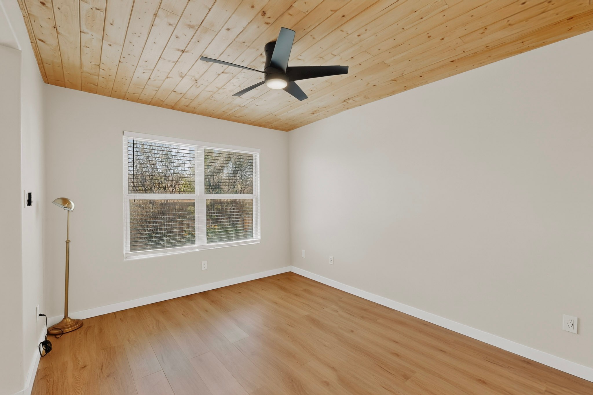 Empty room with wood floors and plank ceiling, black ceiling fan, and window overlooking trees.