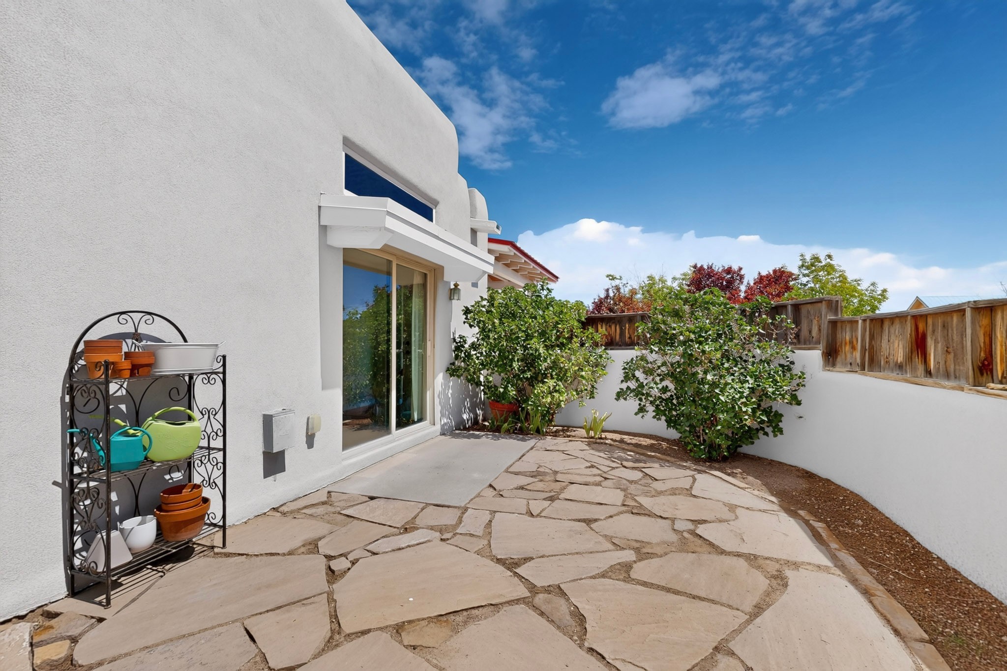 Flagstone patio beside a white stucco home, bordered by shrubs and a wooden privacy fence.