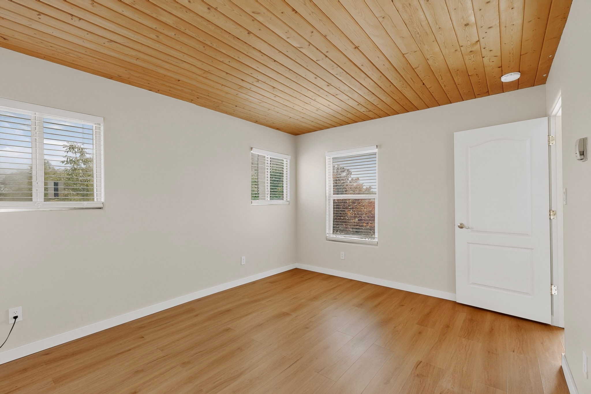 Empty room with wood plank ceiling and flooring, three windows, and a white door.