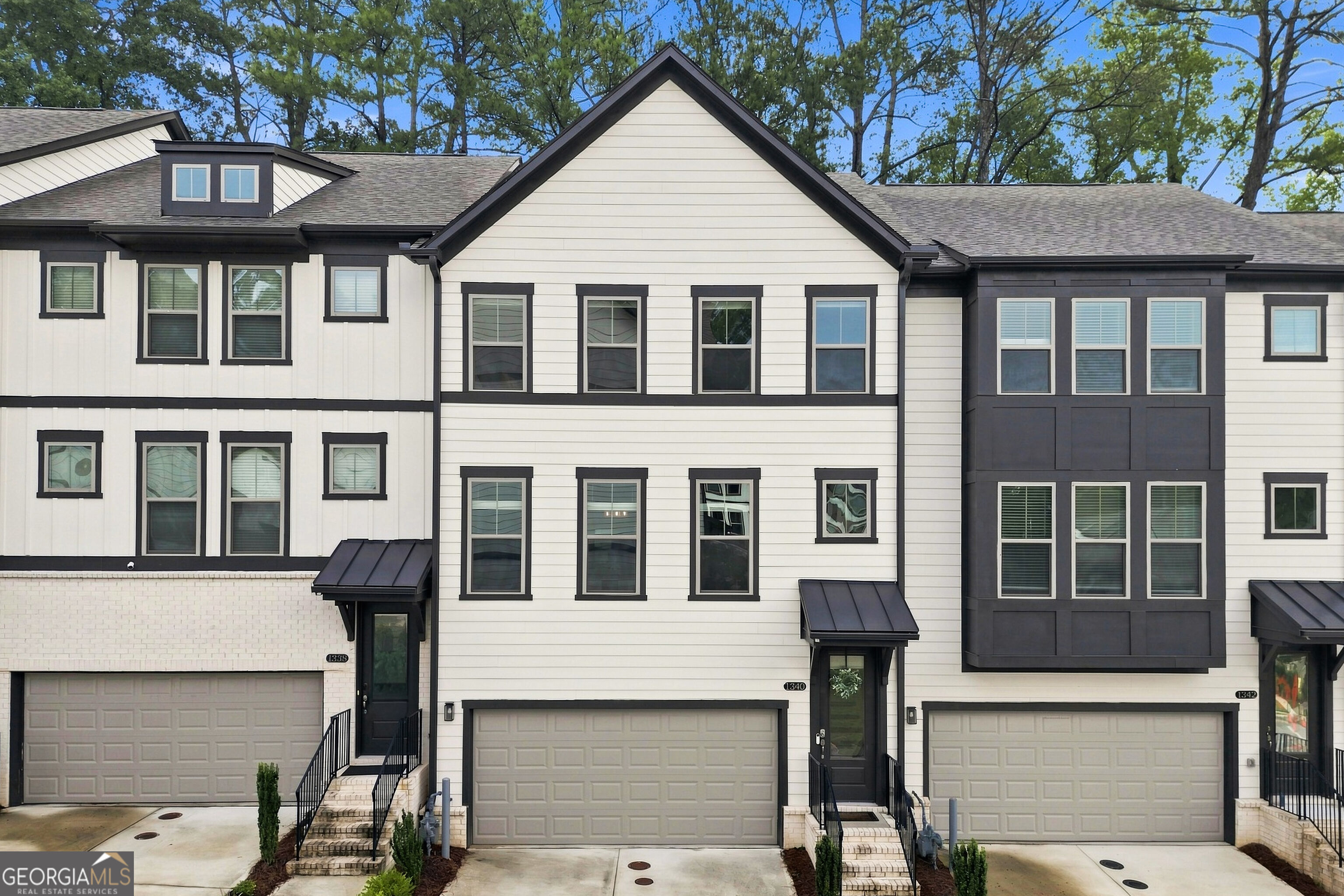 Attached white townhomes with dark trim, front-facing garages, and mature trees behind them.