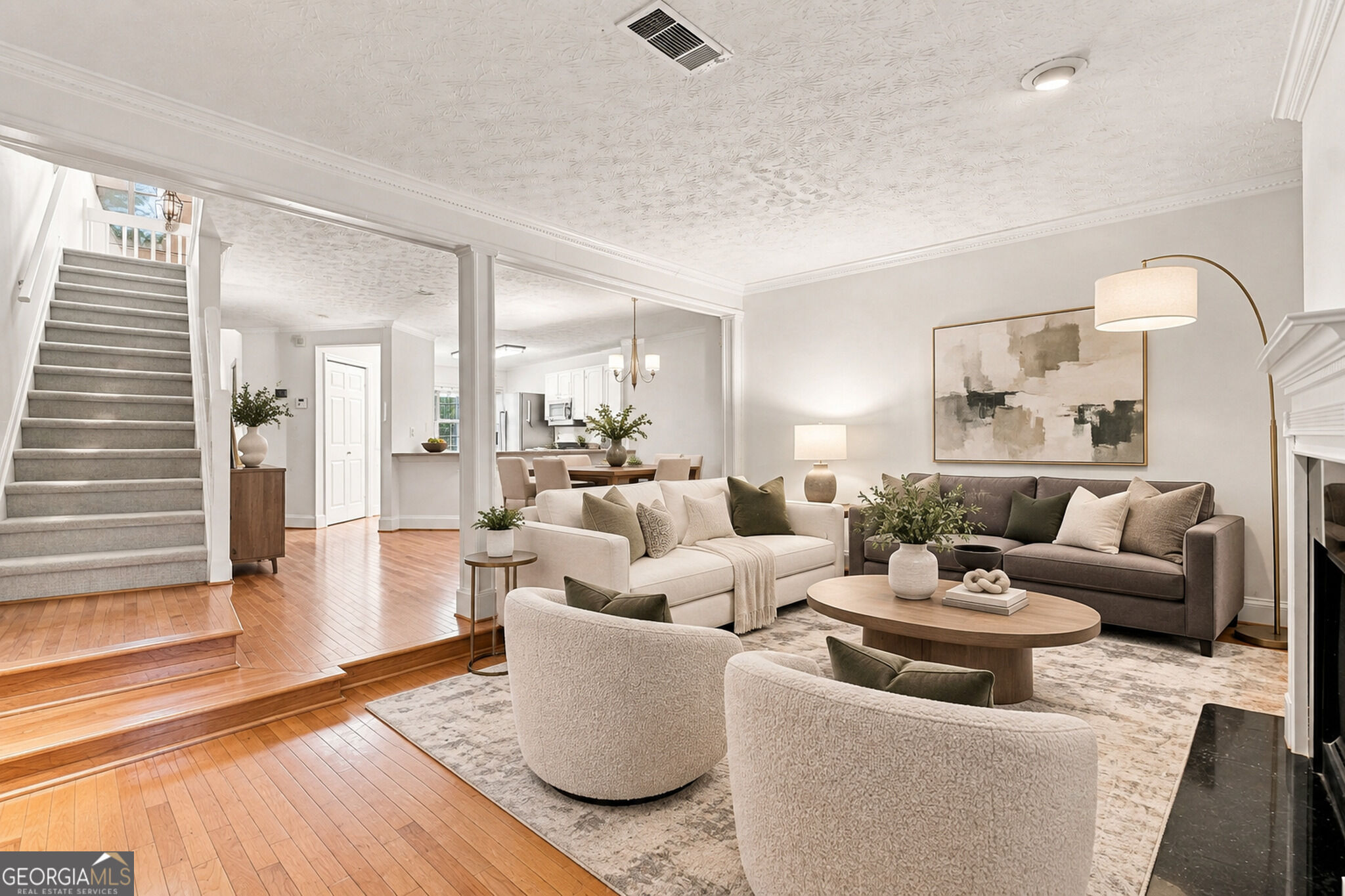 Staged living room with two sofas, rounded chairs, hardwood floors, and a carpeted staircase.