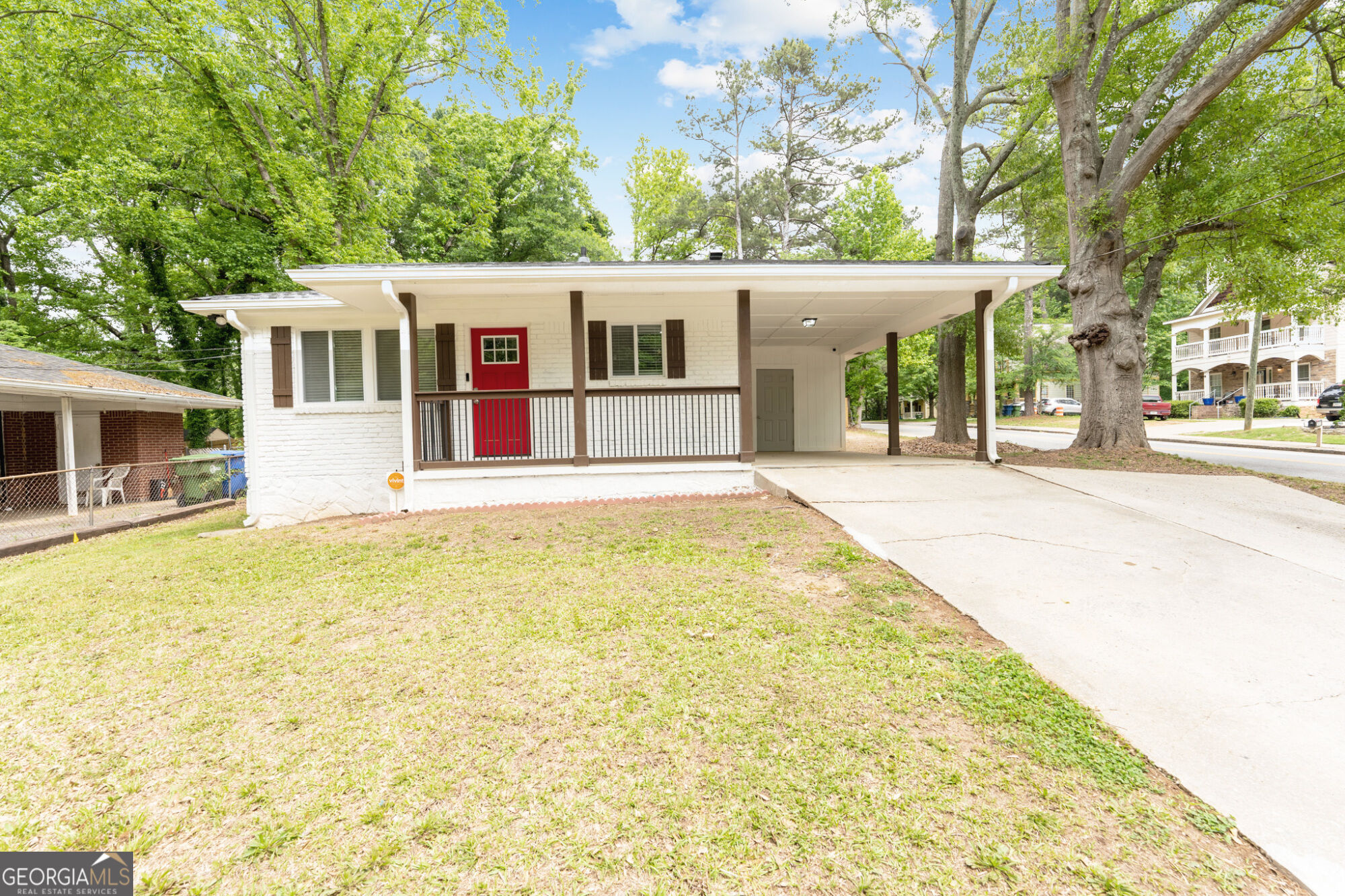 Single-story white brick house with a red front door, covered porch, and wide driveway beneath mature trees.