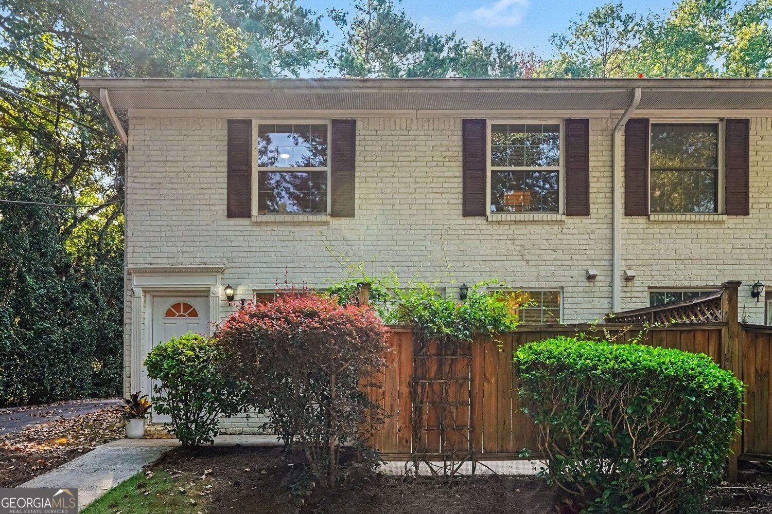 Two-story light brick home with dark shutters, wooden privacy fence, and landscaped shrubs.