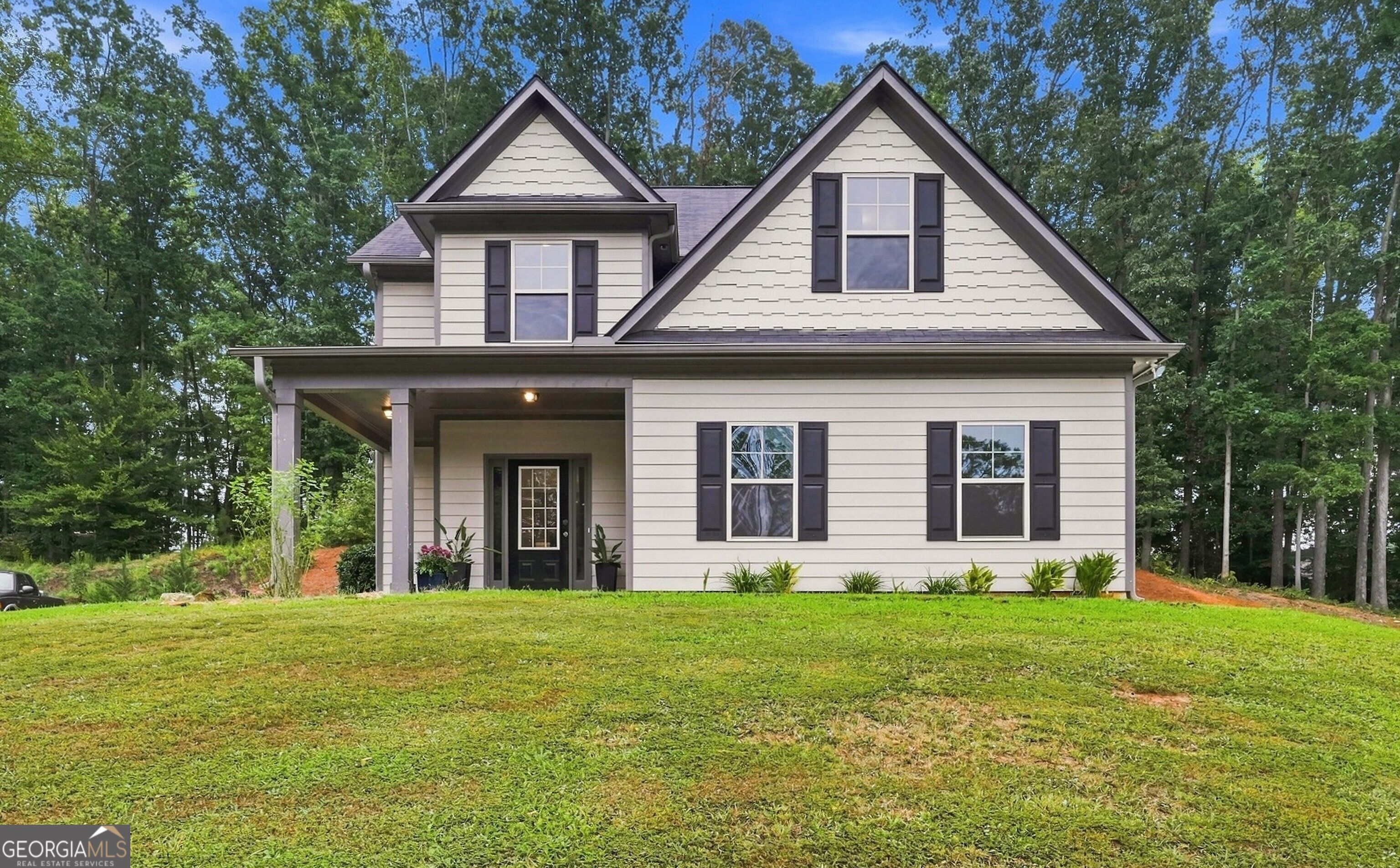 Two-story light-colored house with dark shutters and a covered front porch, surrounded by trees.
