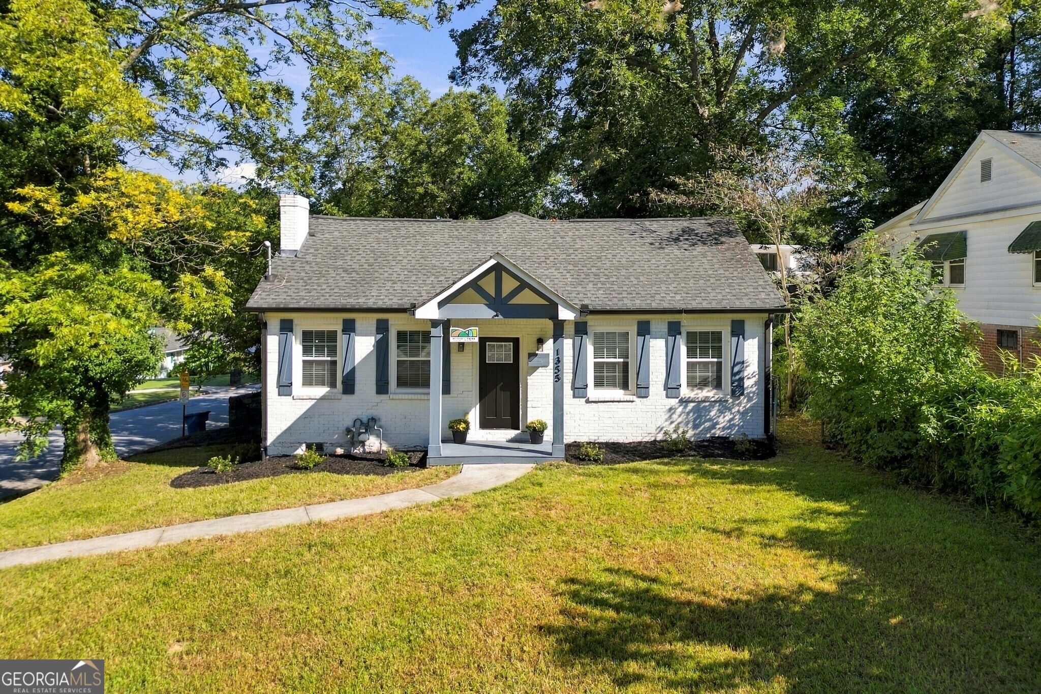 White brick house with blue shutters and a covered entry, framed by mature trees.