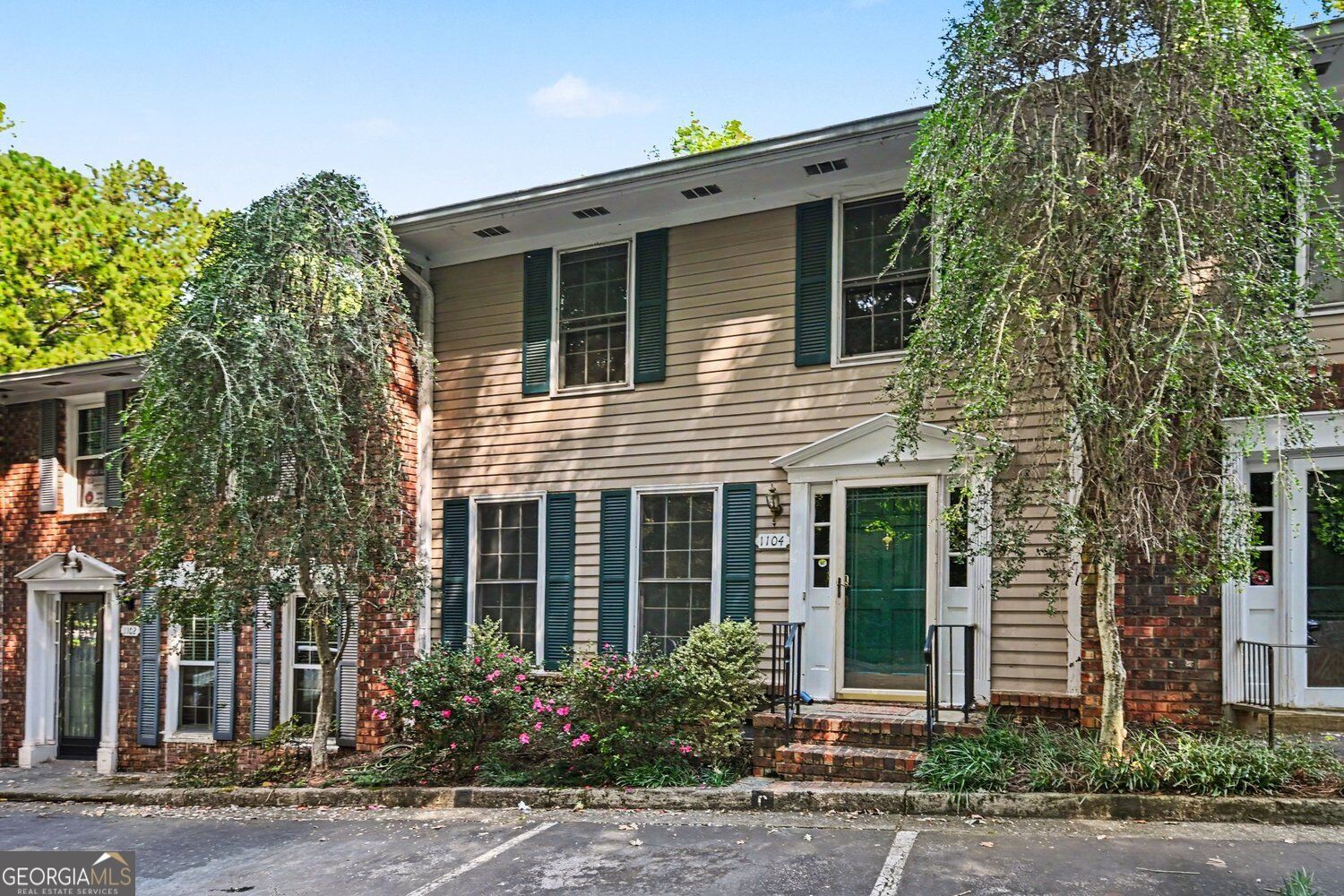 Two-story beige-sided home with a green front door, brick steps, and flowering shrubs.